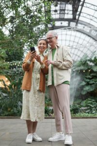 An older couple touring a conservatory, smiling and looking at a smartphone together, surrounded by lush plants. The image illustrates technology enhancing travel experiences for all ages. Photo by Marcus Aurelius on Pexels.