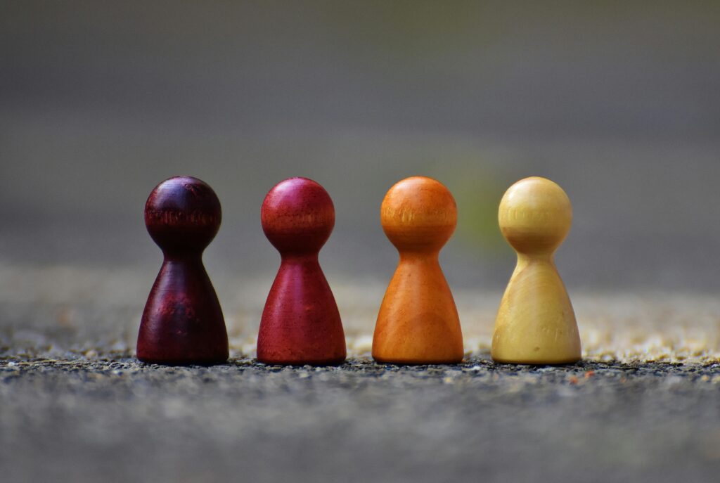 The image shows four wooden game pieces standing in a row, each a different shade of brown, ranging from dark to light. The pieces are arranged in a gradient, symbolizing diversity and inclusion. The background is blurred, drawing focus to the game pieces and their subtle differences in color and texture.